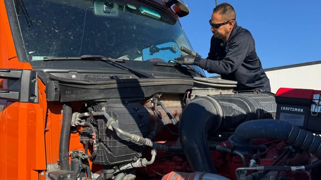 A mechanic in a black uniform replaces a windshield wiper blade on an orange truck as part of a windshield damage checklist.