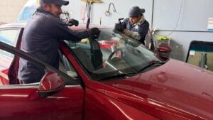 Two people wearing gloves work together to replace or repair the windshield of a red car inside an automotive workshop, providing a practical guide for anyone interested in understanding windshield replacement cost in Utah.