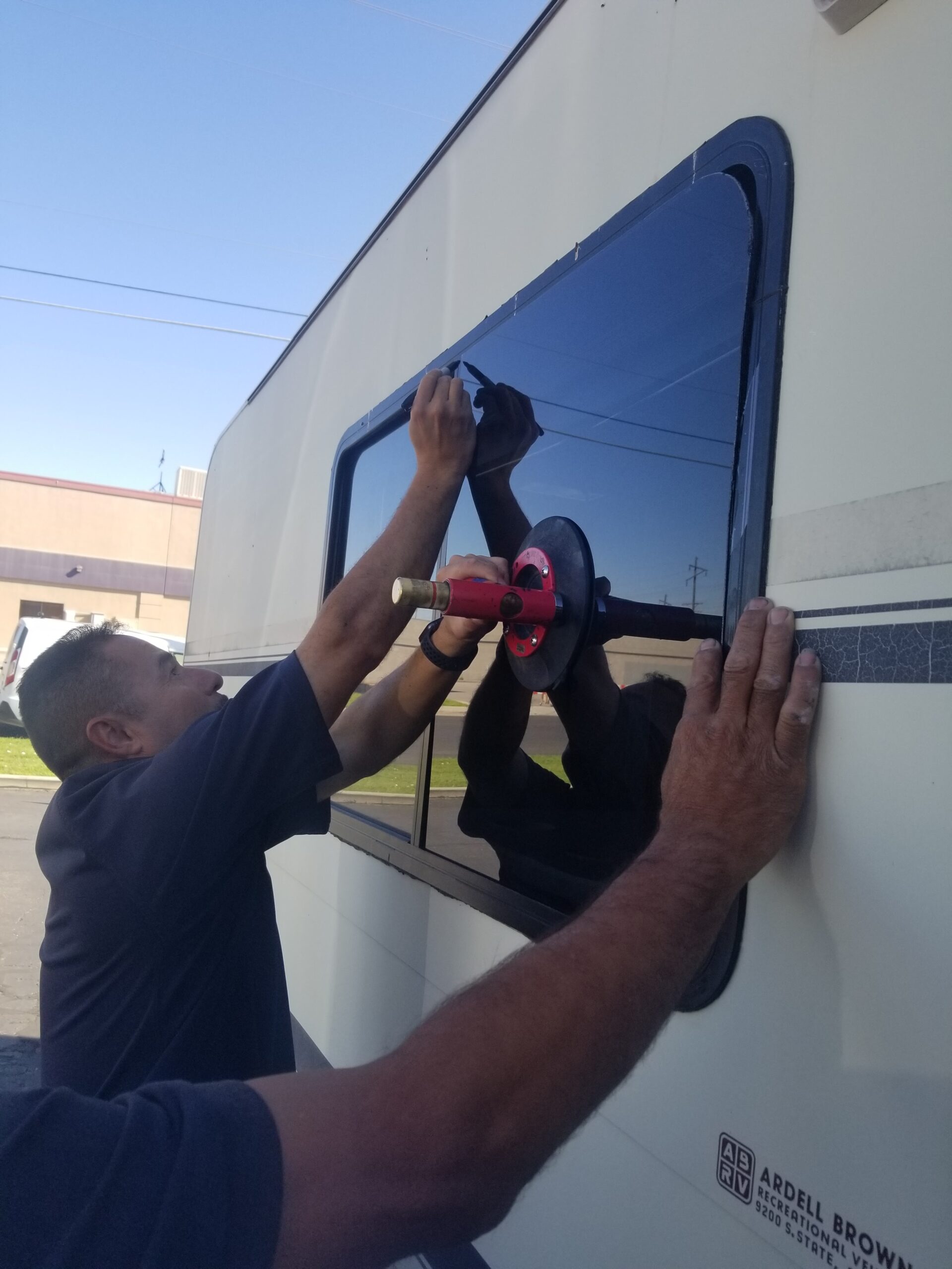 Two people install or adjust a window on the side of a recreational vehicle using a red suction tool and their hands for alignment.
