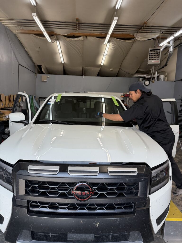 A man wearing a black shirt and cap works on the windshield of a white Nissan truck inside a garage, expertly performing a windshield repair on the vehicle.