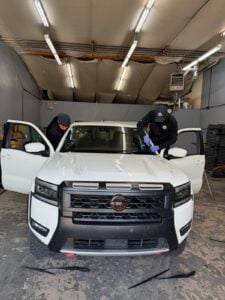 Two people clean the interior of a white Nissan Frontier parked inside a garage with overhead lights and open front doors, preparing the vehicle for windshield repair.