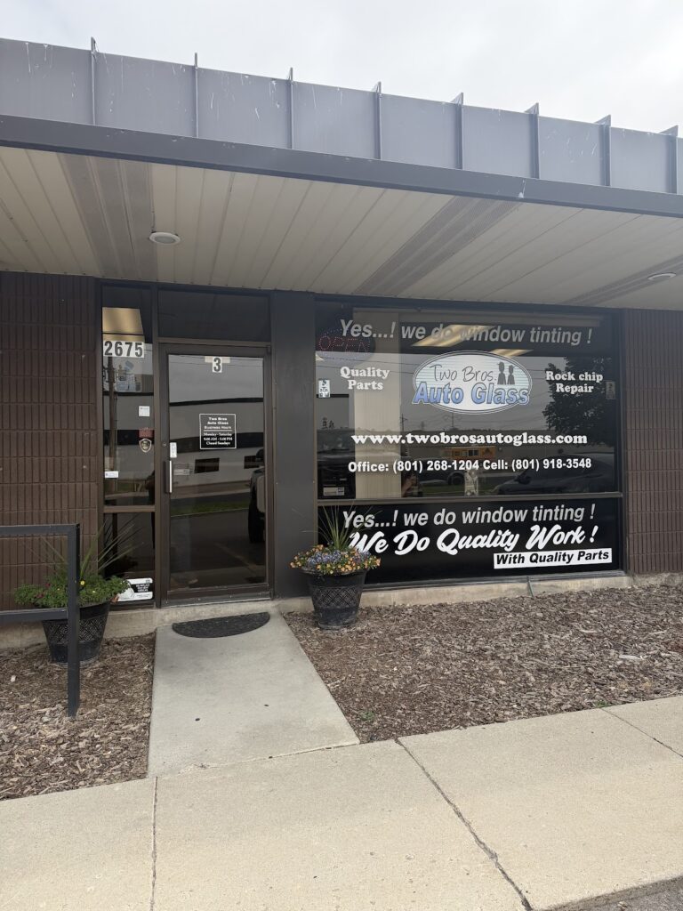 Exterior of Two Bros. II Auto Glass shop with open door, large window sign advertising vehicle windshield repair and replacement, and two potted plants by the entrance.