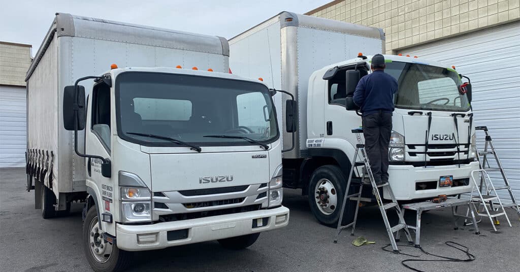 Two white Isuzu box trucks parked side by side, with a person standing on a ladder performing fleet windshield replacement on the passenger side door of the truck on the right.