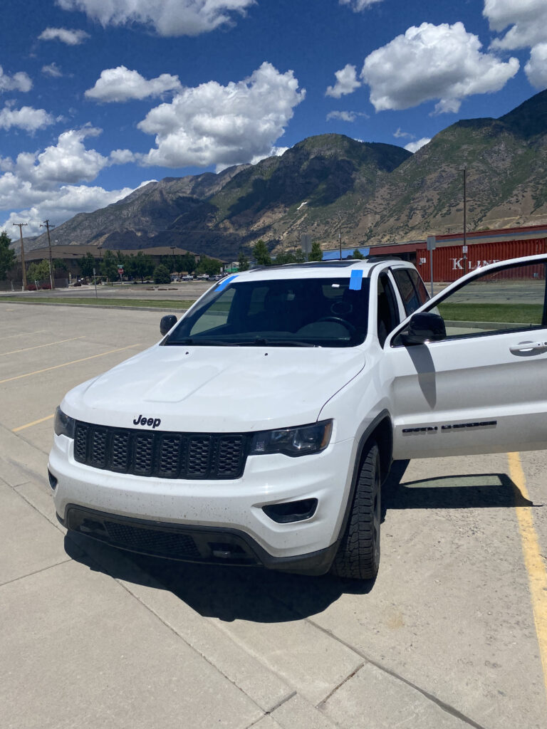A white Jeep Grand Cherokee with an open driver-side door parked in a lot with mountains in the background, perfect for Salt Lake City auto glass services.