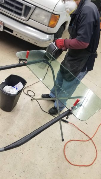 Person wearing gloves and mask works on a large piece of glass clamped to a stand, with a white truck and a trash bin nearby.