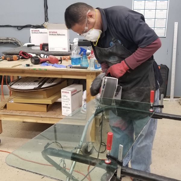 A person wearing safety gear uses a power tool on a glass panel clamped to a workbench in a workshop.