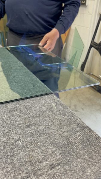 A person holds a thin sheet of clear glass over a padded work surface in a workshop setting.
