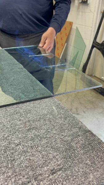Person holding a clear sheet of glass above a gray felt-covered worktable in a workshop setting.