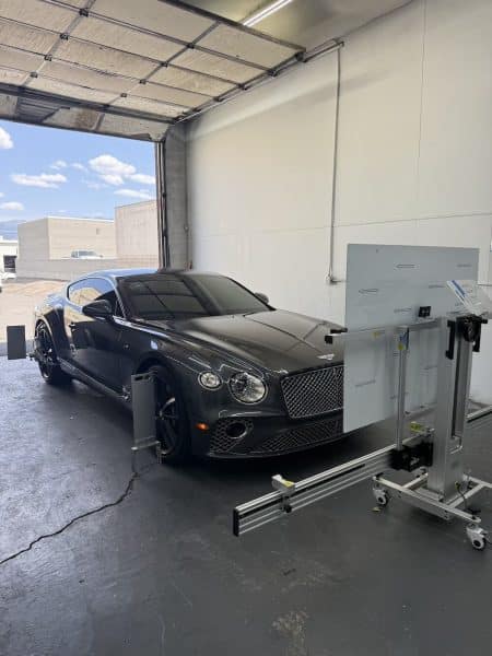 A black luxury car is positioned inside a garage next to a calibration device, with the garage door open to the sky and parking lot, ready for mobile ADAS calibration.